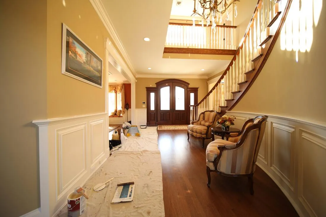 Two-story foyer during interior painting project, drop cloths protecting hardwood floors, wainscoting panels being painted